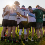 Backs of team members standing in a huddle with their arms around each other and sunlight shining above.