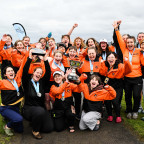 Women's rowing team wearing L&C orange jackets and cheering with their trophy.