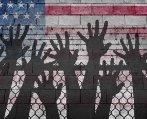 Shadow of hands reaching up over a chain link fence with an American flag as the background.